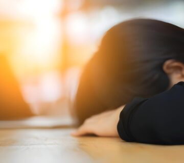 A tired person with dark hair rests their head on a wooden desk with a blurred laptop in the background