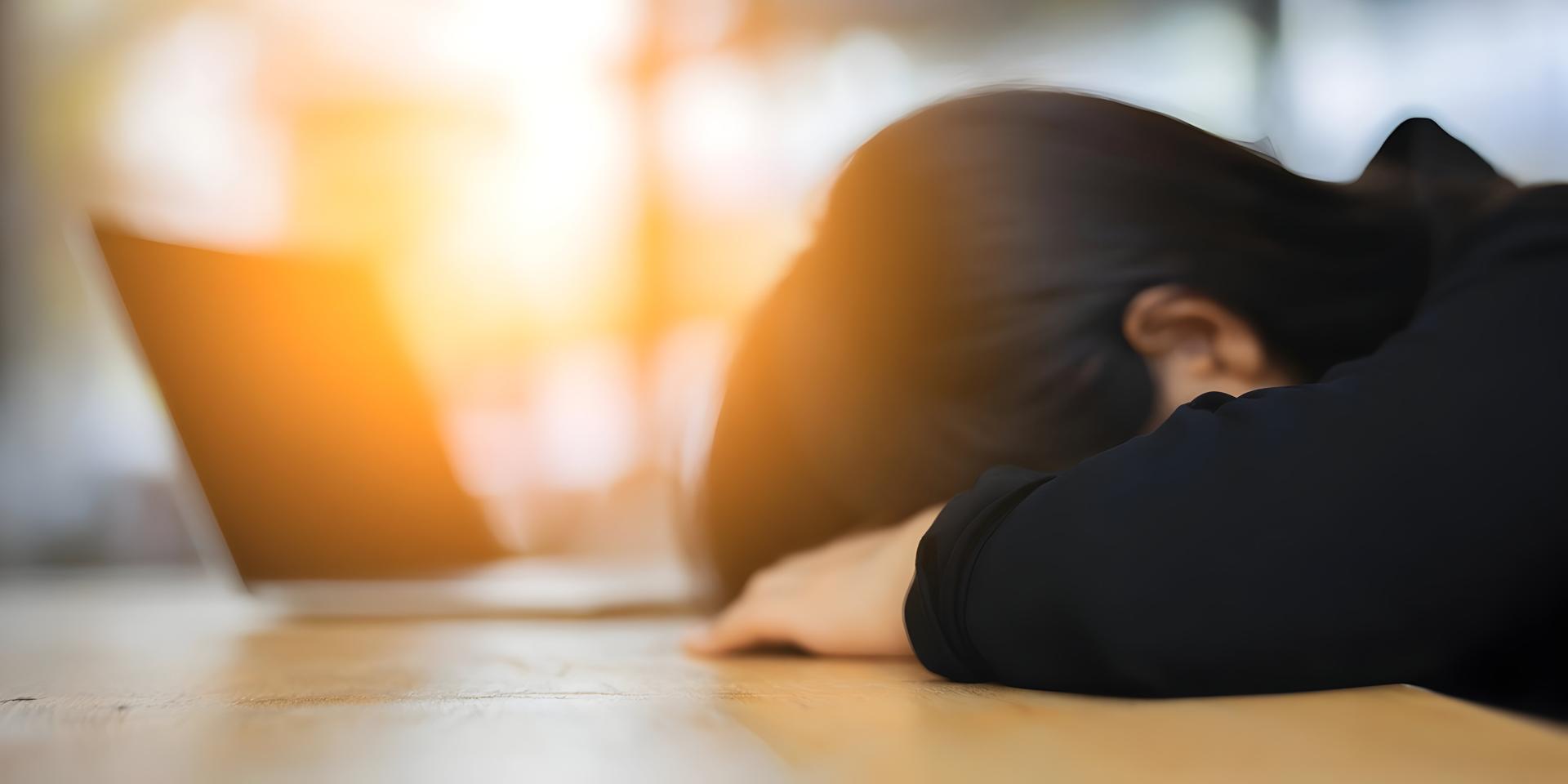 A tired person with dark hair rests their head on a wooden desk with a blurred laptop in the background